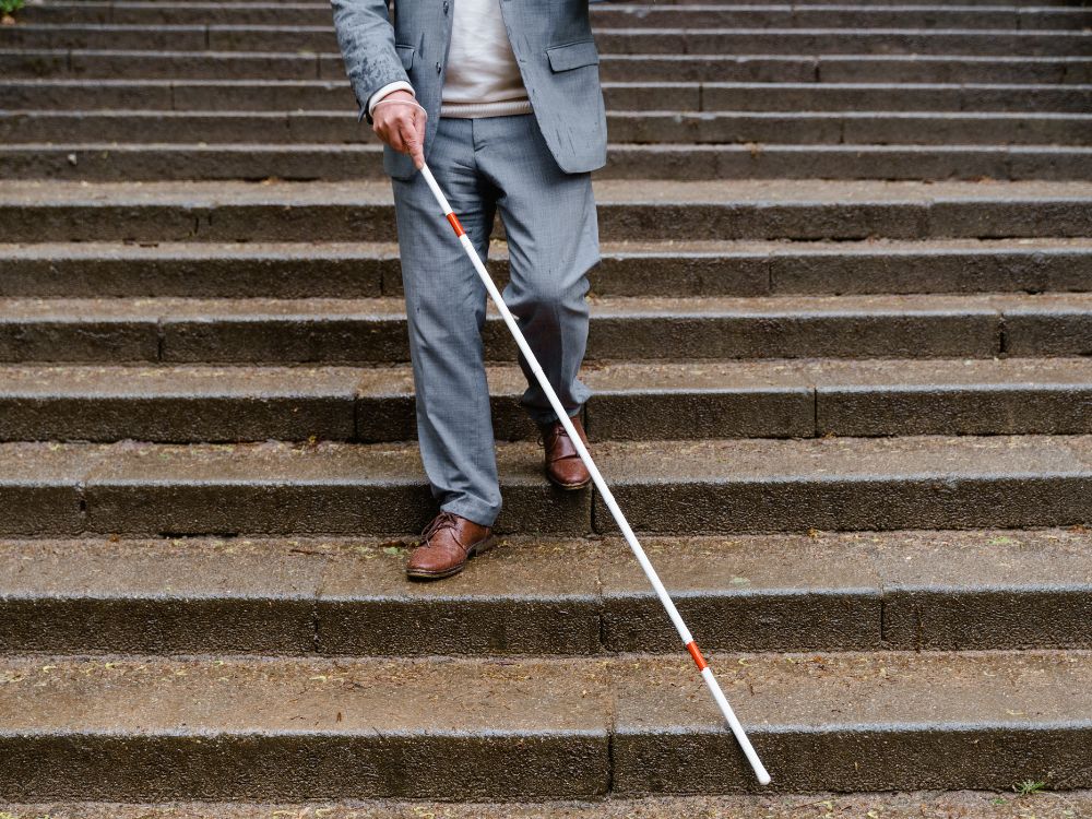 Homem com deficiência visual desce escada usando bengala, ilustrando a falta de acessibilidade em espaços públicos e barreiras arquitetônicas.