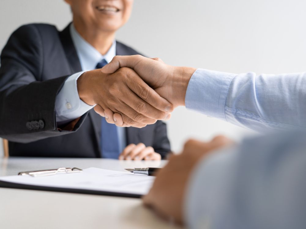 Pessoa de terno cumprimenta um cliente com um aperto de mão sobre a mesa, simbolizando a contratação de um advogado particular de confiança.