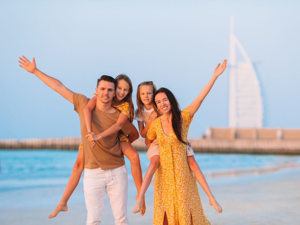 Família sorridente curtindo as férias na praia ao pôr do sol, com crianças nos ombros e pais felizes diante do mar e do Burj Al Arab ao fundo.