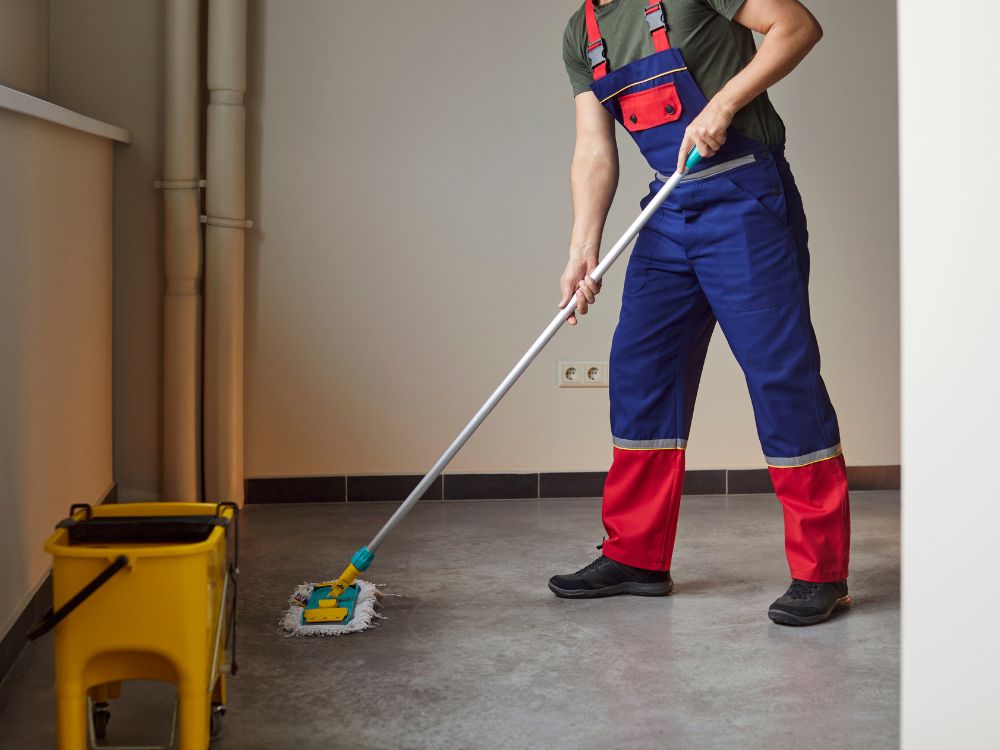 Profissional limpando o chão com mop ao lado de balde amarelo, representando manutenção do prédio: quem paga, inquilino ou proprietário.